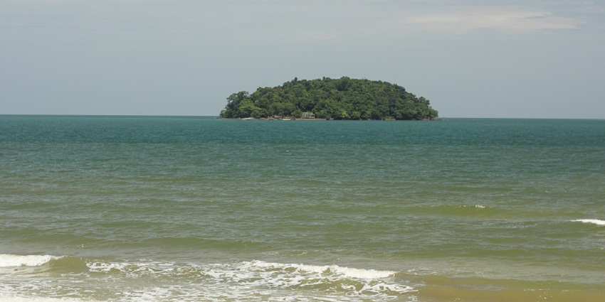 A small, lush green island sits alone in the calm sea under a cloudy sky, representing the natural beauty of the islands in Cambodia.