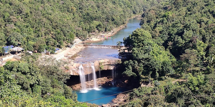 Krang Suri falls from above with clear blue water body, distant people and greenery.
