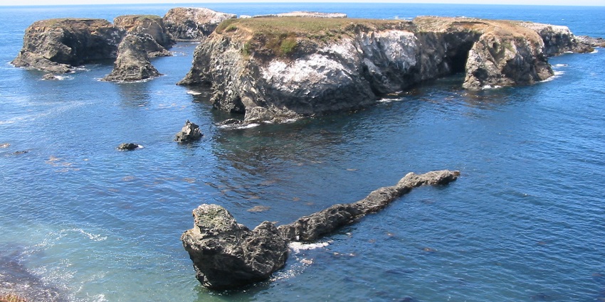 Jagged coastal rocks into the clear blue sea, surrounded by cliffs and small waves.