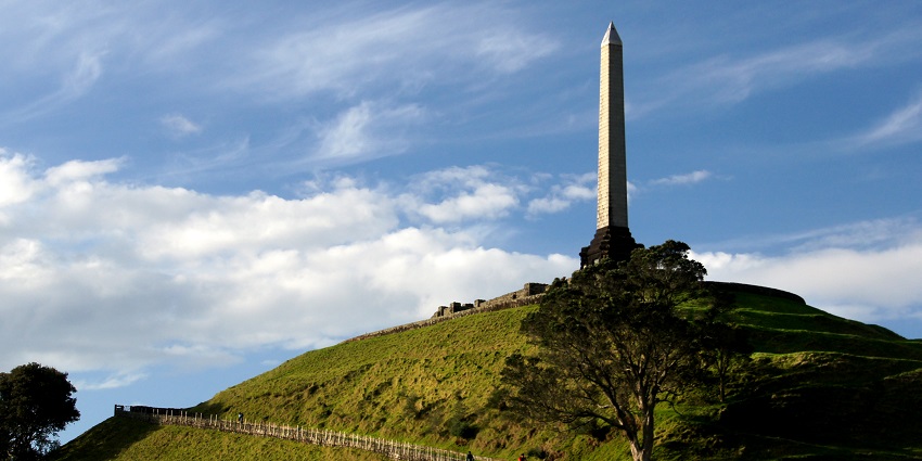A tall obelisk perched atop a grassy volcanic hill, one of the historic monuments in New Zealand, surrounded by a winding road and lush greenery.