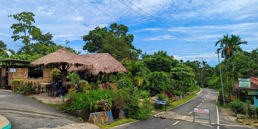 Street in Mawlynnong village with distant houses, multiple sign boards and greenery.
