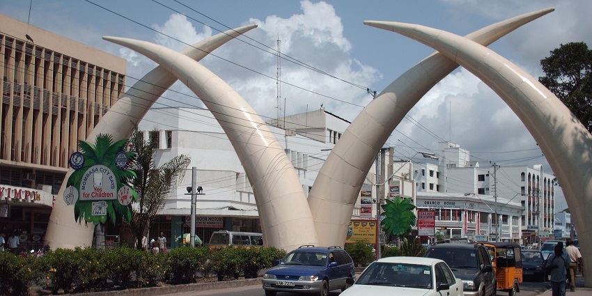 Mombasa Tusks of Kenya arching over a busy road filled with cars and daily city traffic.