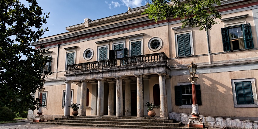 Historic neoclassical mansion with columns, green shutters, and stone steps under blue sky.