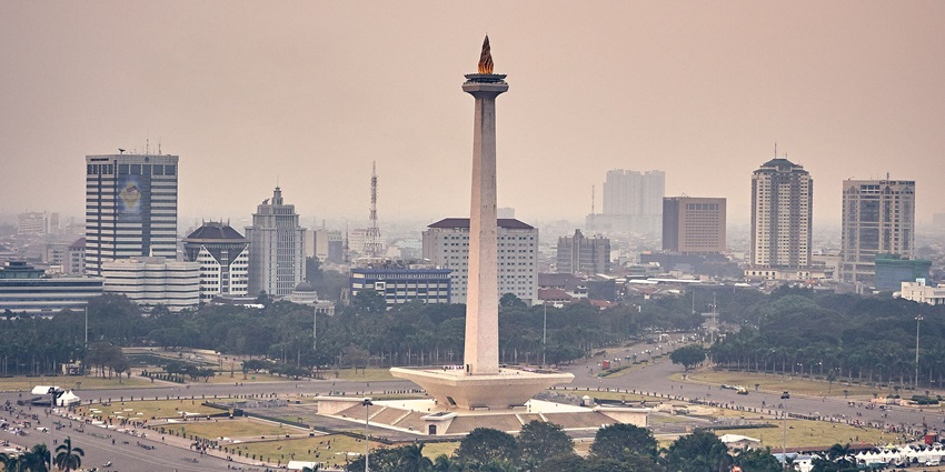 A view of the National Monument (Monas) in Jakarta, Indonesia, standing tall with a golden flame at its peak, surrounded by modern high-rise buildings and a large public square with trees and pathways.