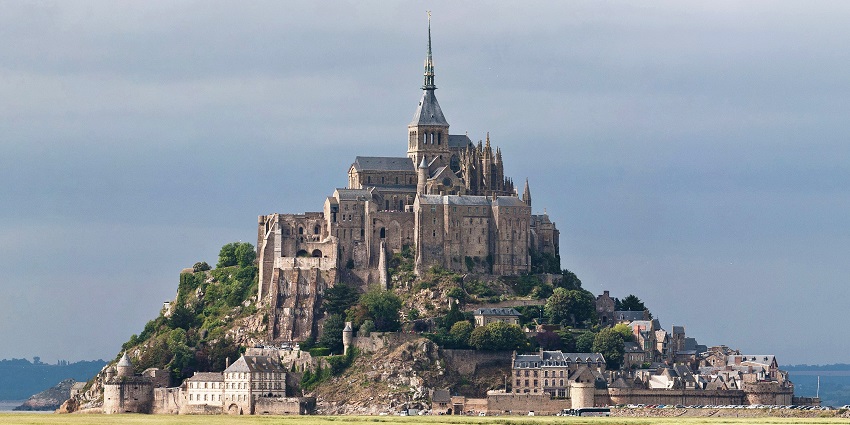 The Mont Saint-Michel abbey stands on a rocky island under the clear sky with lush greenery around.