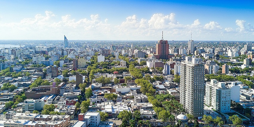 Aerial view of a modern city with a mix of high-rise and low-rise buildings, green trees, and a clear blue sky.