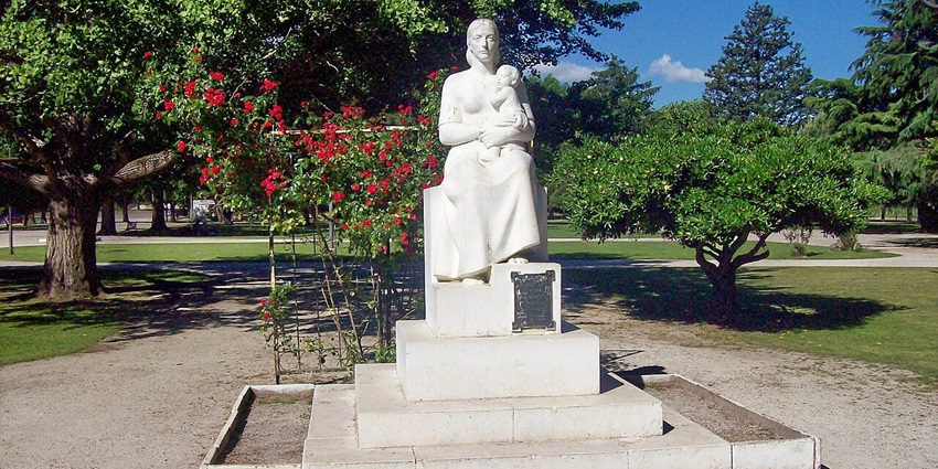 A white statue of a woman holding a baby, signage on the platform of the statue, and flowers