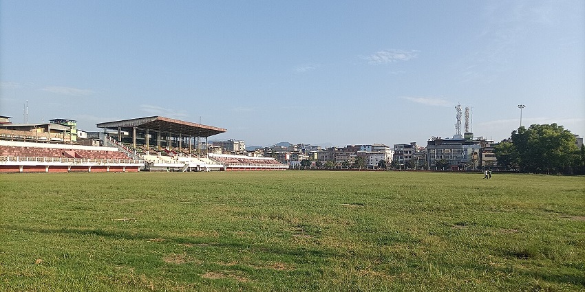 Polo ground with green grass plains, distant buildings, a sitting place for people.