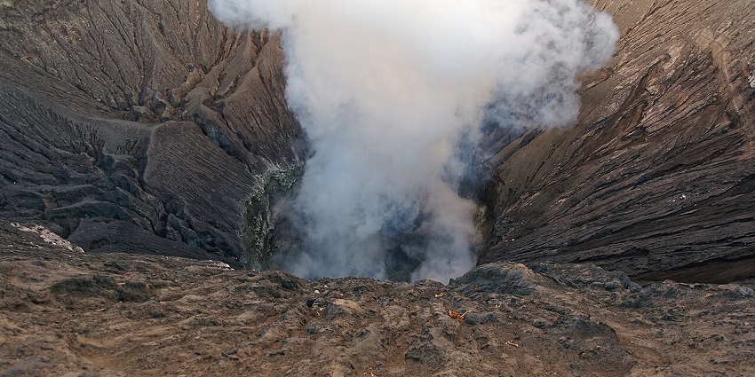 A stunning landscape of volcanic mountains shrouded in mist, with one actively emitting a plume of white smoke into the clear morning sky.
