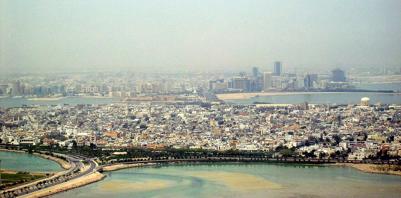 A scenic aerial view of Muharraq and Manama in Bahrain, showing buildings, bridges, and coastline under a clear sky.