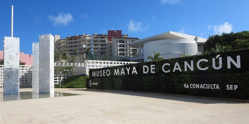 Entrance of Museo Maya de Cancún with modern sculptures and buildings under a blue sky.