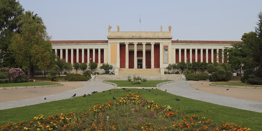 Neoclassical building with columns, statues on the roof, and a colourful flower garden in front.