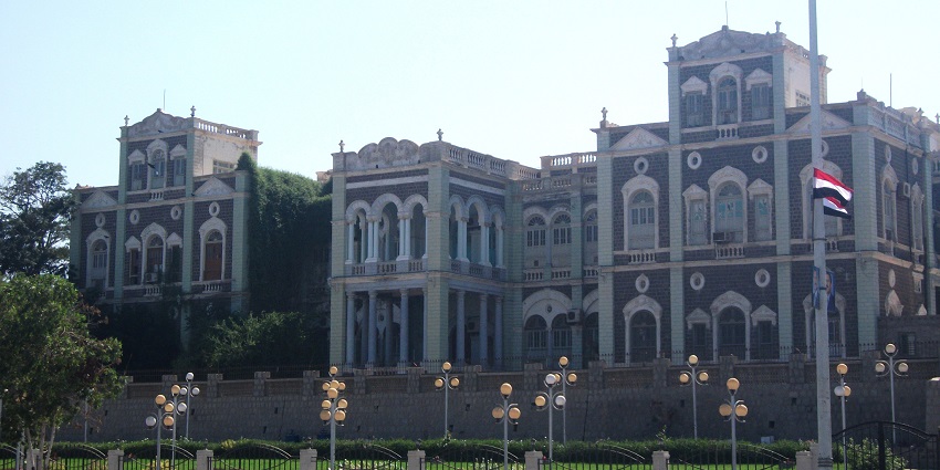 Large white building with arched windows, tall palm trees, one of the best historical places in Yemen.