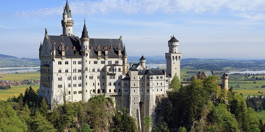 Neuschwanstein Castle is perched on a forested hill with fields and mountains beyond, one of the most beautiful historical places in Europe.
