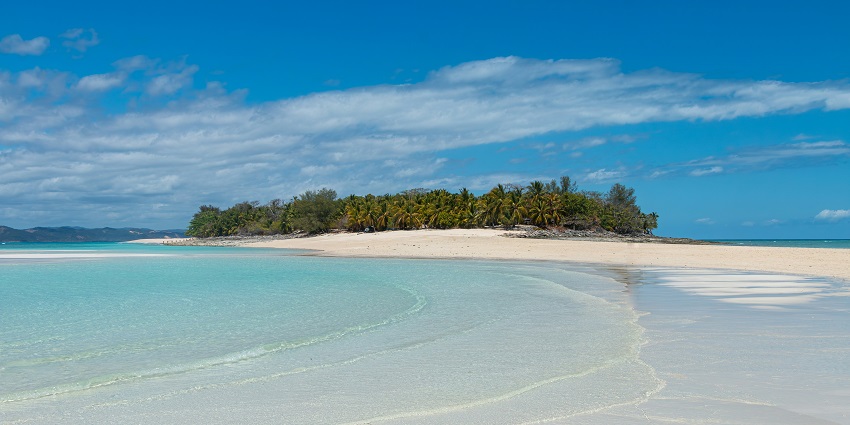 A tropical island with white sandy beach, clear blue water, and green hills under a bright sunny sky.
