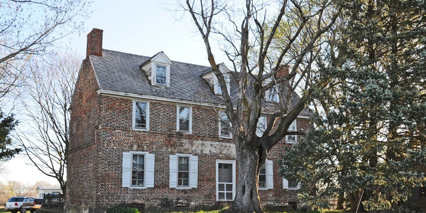 Historic red-brick colonial house with shuttered windows and large trees in the front yard.