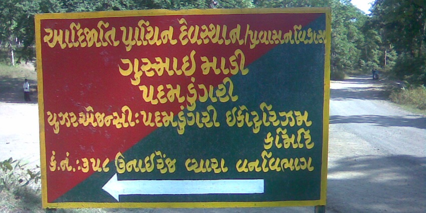 A Gujarati signboard in a forested area directing visitors to a local temple or site of interest.