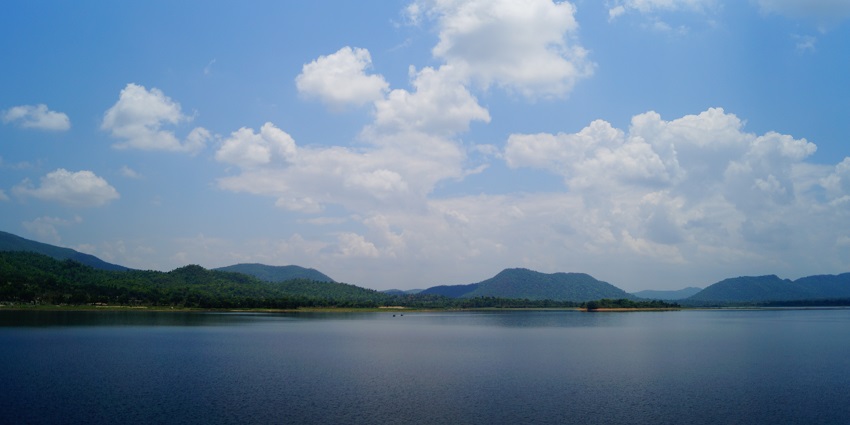 Glide over the tranquil waters of Dimna Lake, surrounded by wooded hills and clear skies.
