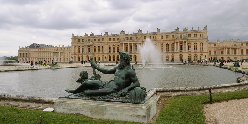 Fountain with statues in front of the Grand Palace of Versailles under a cloudy sky, one of the top historical places in France.