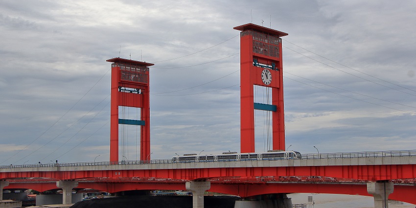 Red vertical-lift bridge with clock towers spanning a wide brown river under a cloudy sky.