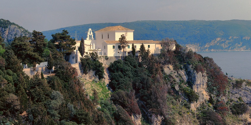 A scenic view of a monastery perched on a forested cliff overlooking the sea and hills.