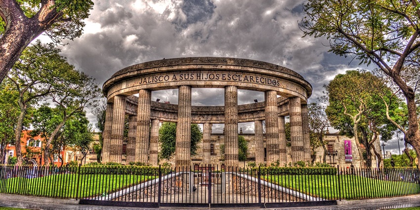 Rounded stone rotunda with columns and trees, honouring local heroes in a quiet plaza.