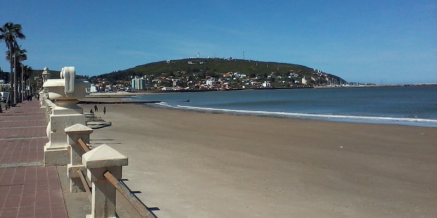 A scenic beach with a paved promenade and a hillside town in the background highlights a coastal place to visit in Uruguay.