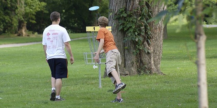 People playing disc golf in a grassy park, aiming a disc toward the metal basket under a blue sky.