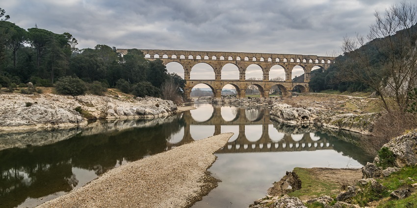 An ancient multi-level stone aqueduct reflected in a calm river, surrounded by rocky banks.