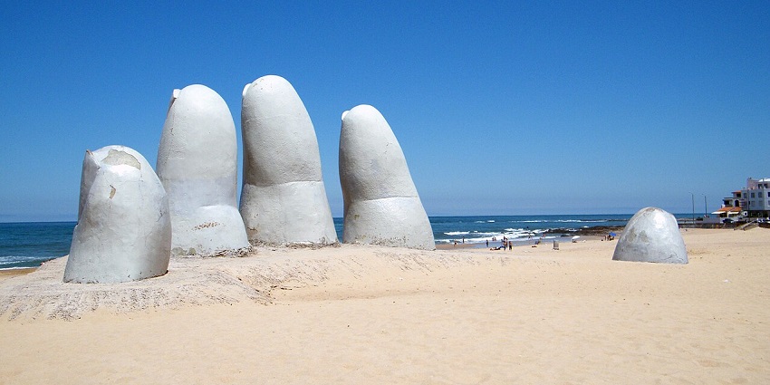 Large sculpted fingers emerging from the sand on a beach with a clear blue sky and ocean waves in the background.
