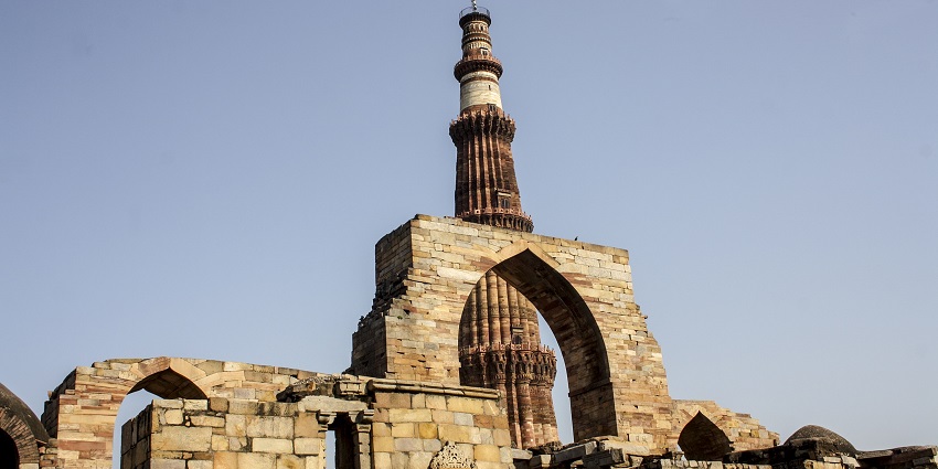 Ancient stone ruins with an arched gateway in front of a tall, historic minaret under a clear sky.