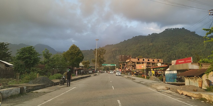 Welcome gate to Roing with police barricades, vehicles, tall green trees and mountains