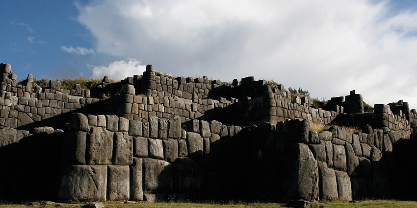 Sacsayhuamán walls with unique rocky architecture, green grass plains, and cloudy sky