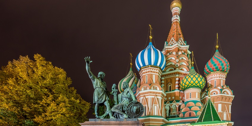 Night view of Saint Basil's Cathedral in Moscow, Russia, with its colorful onion-shaped domes illuminated, and the Monument to Minin and Pozharsky in the foreground, set against a dark sky and a tree with golden leaves.