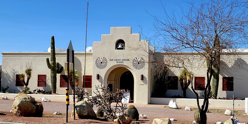 Stucco mission-style school building with arched entry, and cacti desert landscaping.