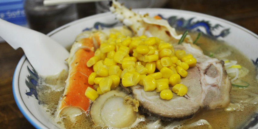 A bowl of miso ramen topped with corn, pork slices, crab legs, and scallops in a rich broth.