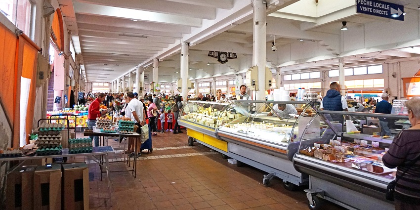 Entrance to Marché Forville showing signboards and people walking near fresh food stalls.