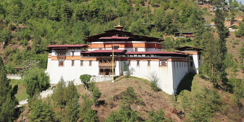 Small fortress-like monastery with stone walls and red detailing, nestled against green hills under a clear sky.