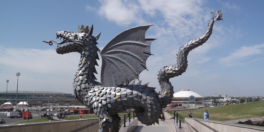 Massive dragon statues surround a round modern building, with visitors walking near sculpture bases.