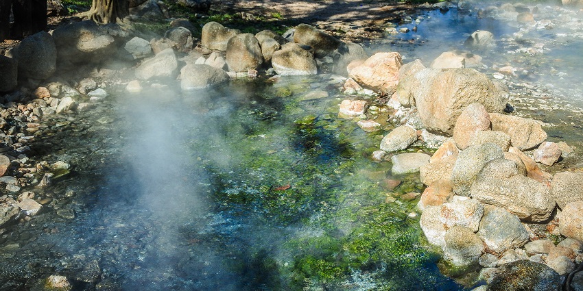 Steam rising from a circular hot spring pool surrounded by stone and green vegetation.