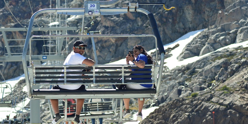 A couple sits on a chairlift ride at Falls Creek, surrounded by snowy mountains, pine trees, and clear blue skies.
