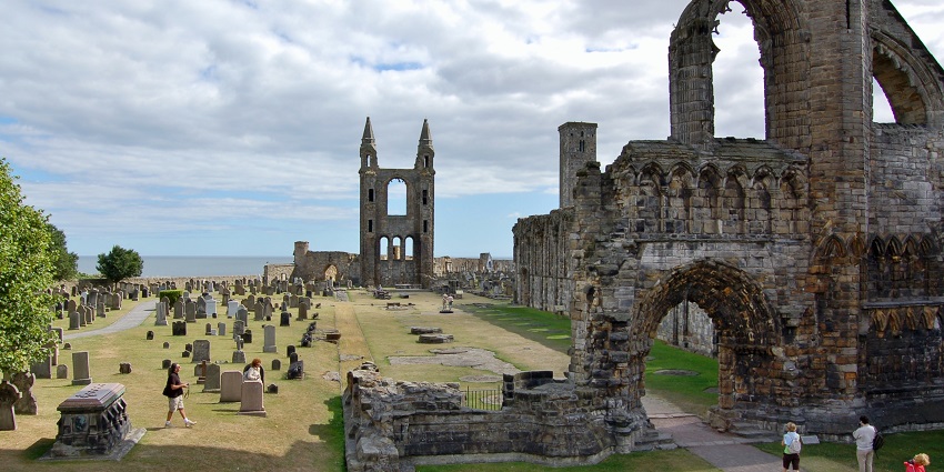 Ruins of an ancient cathedral with twin towers and gravestones scattered with greenery.