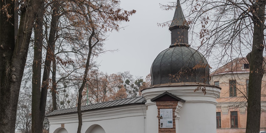 A historic small white chapel with a dark dome roof, surrounded by trees, one of the historical places in Ukraine.