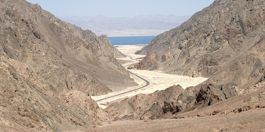 Winding road through rocky desert mountains leading to a distant blue sea under clear sky.