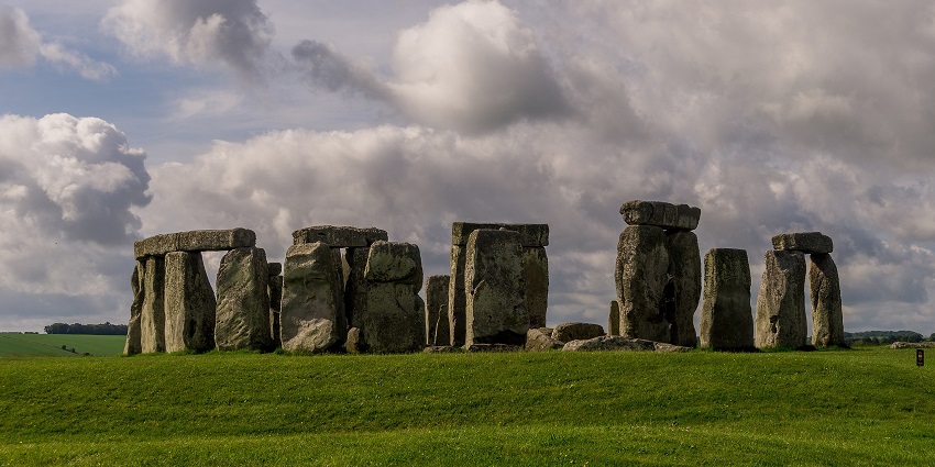 A view of Stonehenge with tall, wide stone pillars and green grass plains under the cloudy sky.