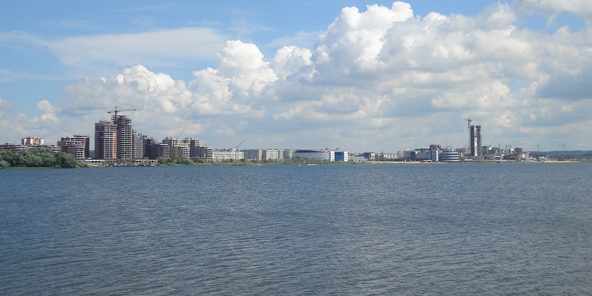 Wide riverside path with benches, public sculptures, and views of Kazan Arena across calm water.