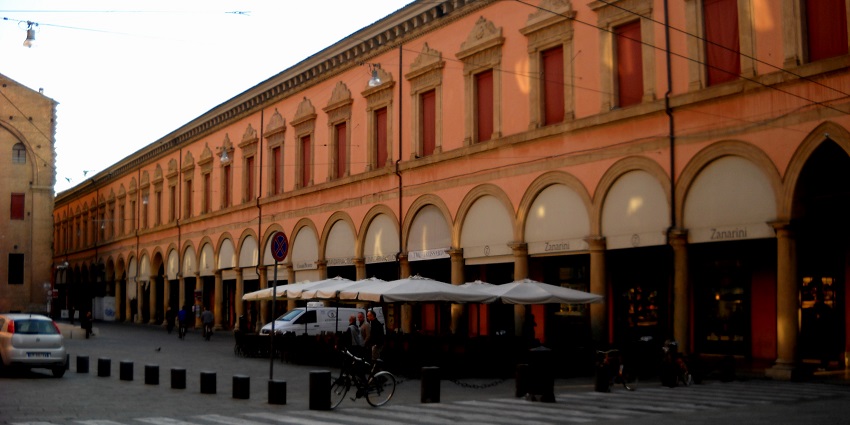 Covered walkway with rows of arches, red-toned columns, and soft light streaming across the corridor.
