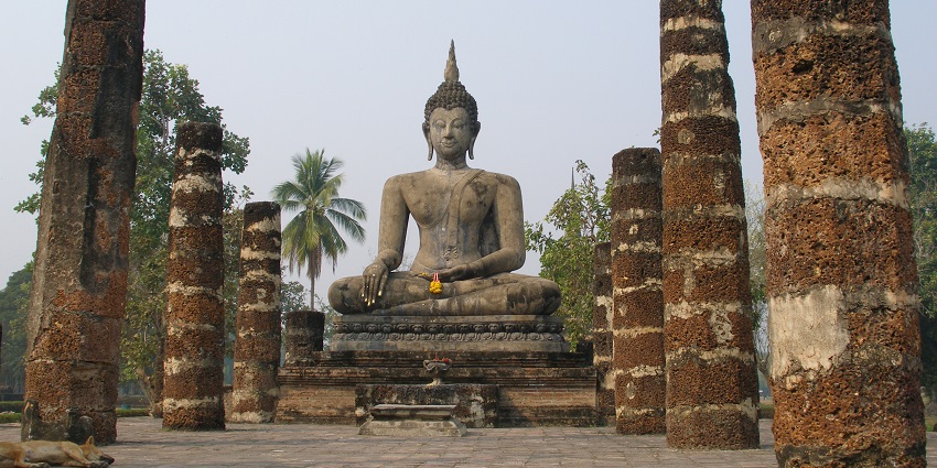 Sukhothai with an old Buddha statue, multiple pillars, and tall green trees in the back.