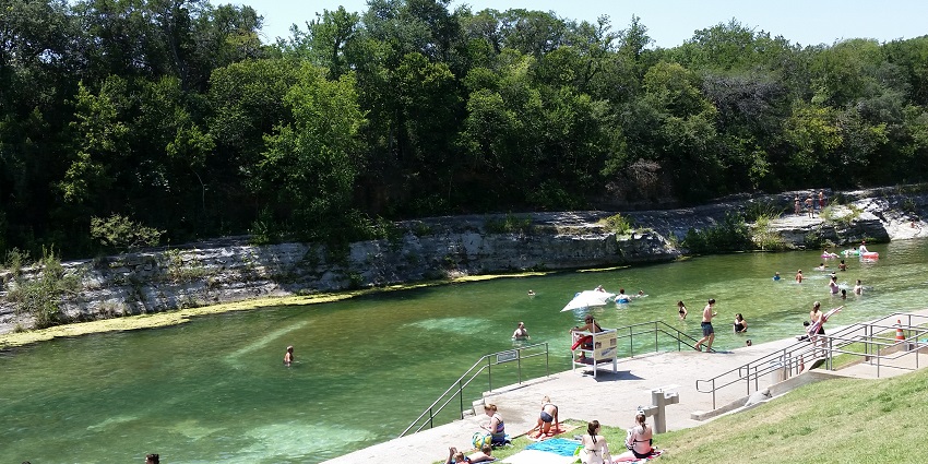 People swimming and lounging on grassy banks beside the calm spring-fed pool