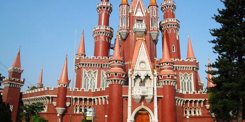 A red-brick castle with tall spires, arched windows, intricate detailing, and a large wooden entrance door.